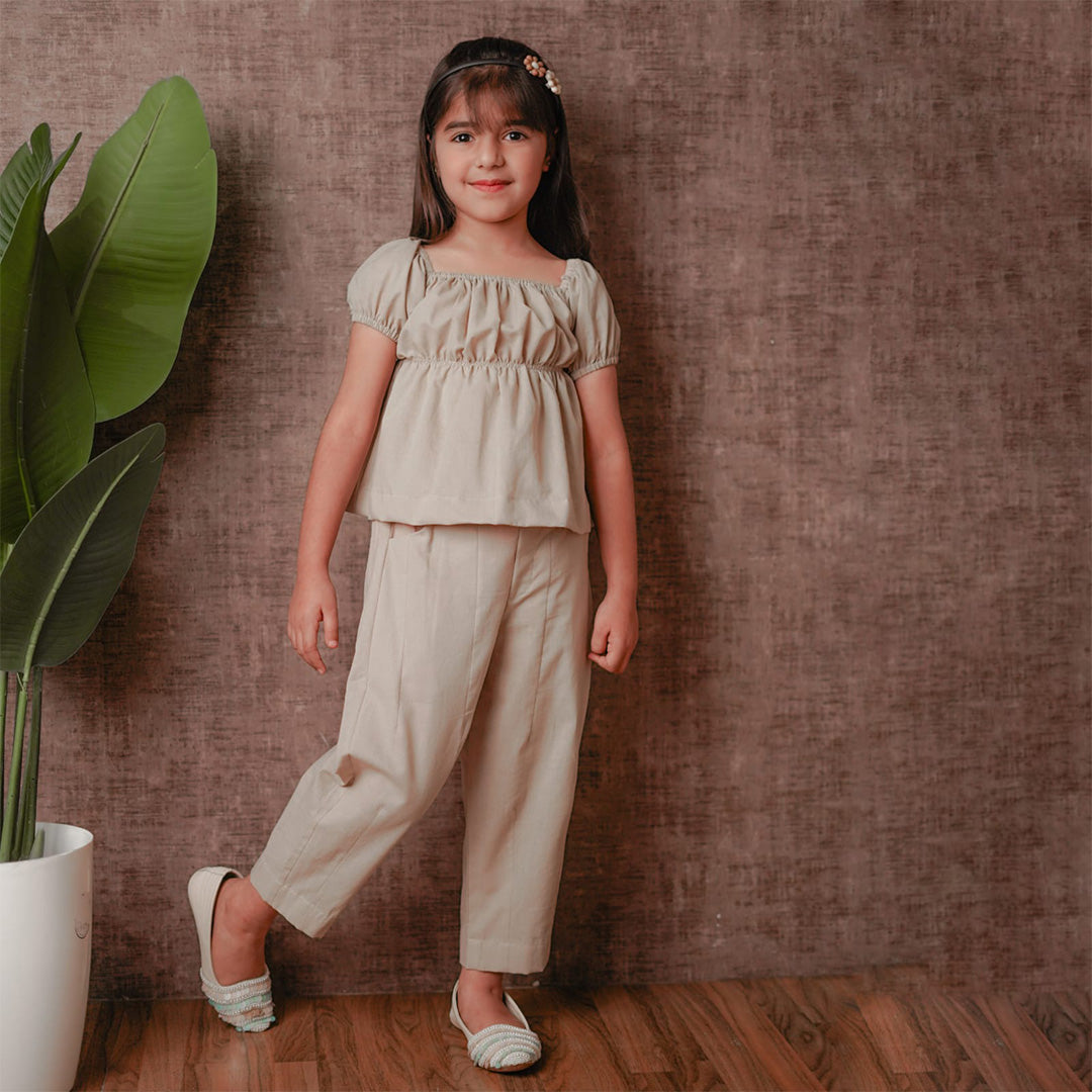 Young girl in a beige outfit standing against a brown textured wall with a plant to her left.