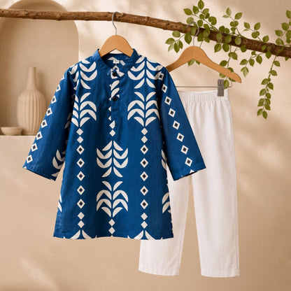 A two-piece children's outfit hanging on wooden hangers, featuring a blue short-sleeve tunic top with a white geometric leaf and diamond pattern, paired with white cropped pants, set against a warm beige background with a decorative branch and green leaves.