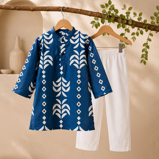A two-piece children's outfit hanging on wooden hangers, featuring a blue short-sleeve tunic top with a white geometric leaf and diamond pattern, paired with white cropped pants, set against a warm beige background with a decorative branch and green leaves.