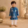 A young boy stands smiling in a studio setting wearing a blue kurta with white block-print style patterns, white pants, and tan shoes. The background features warm beige drapery, large ceramic pots with dried flowers, and soft natural lighting.