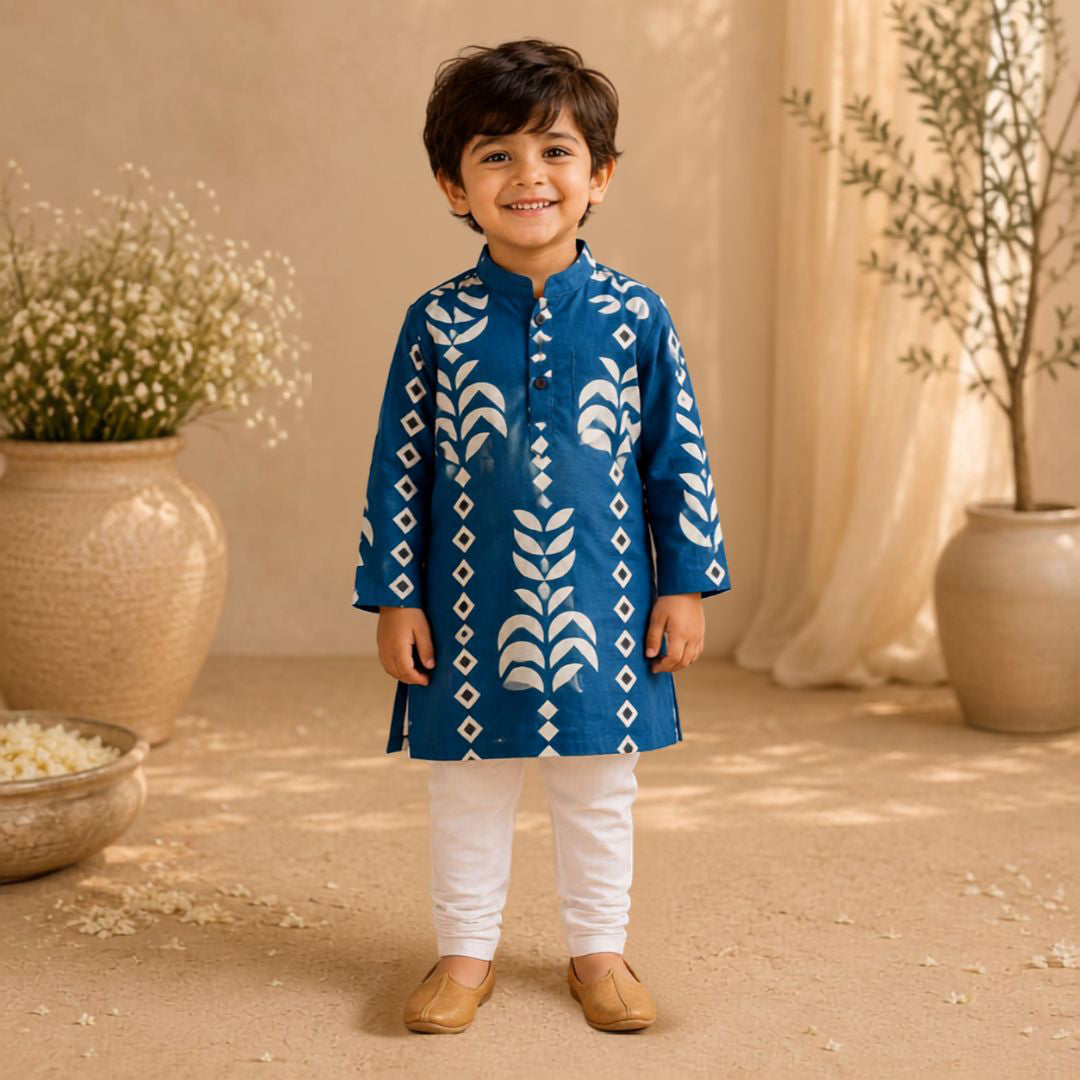 A young boy stands smiling in a studio setting wearing a blue kurta with white block-print style patterns, white pants, and tan shoes. The background features warm beige drapery, large ceramic pots with dried flowers, and soft natural lighting.